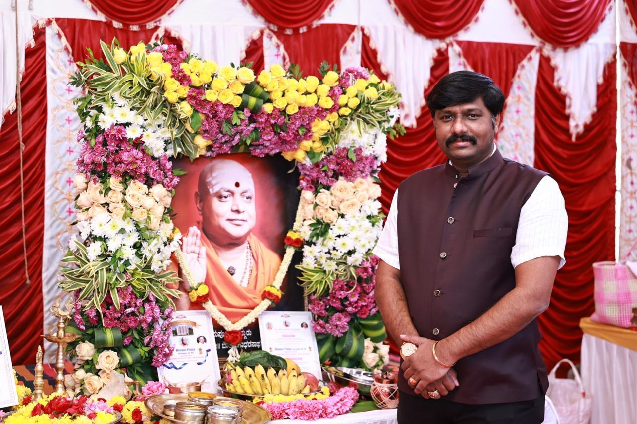 Standing beside the floral-decorated portrait of the seer during the Bhoomi Puja
